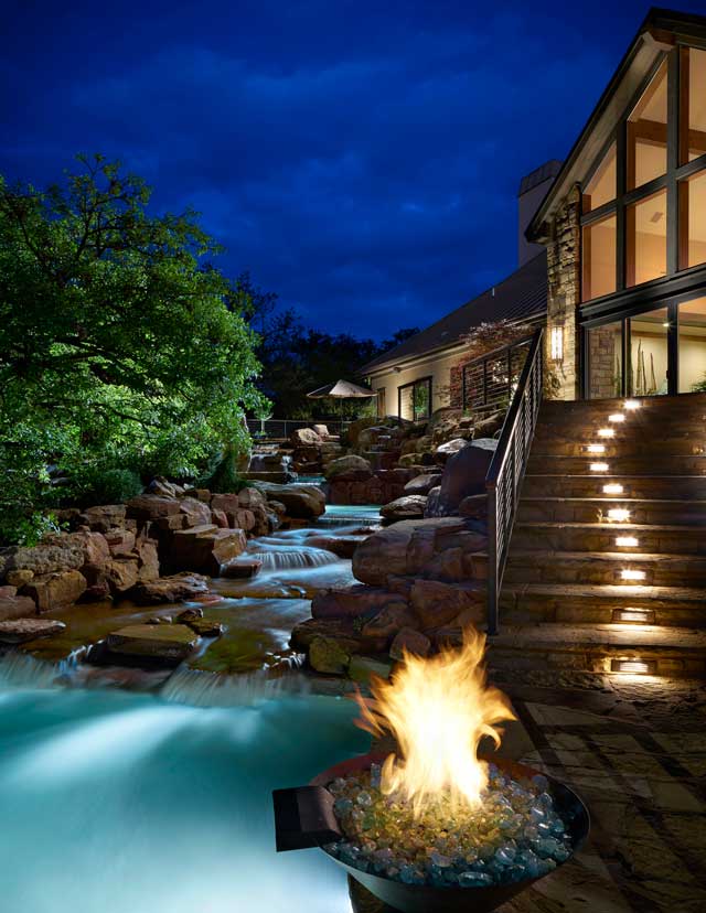 Dramatic lighting illuminates the rear setting of this luxurious home. A natural waterfall cascades over rocks, rushing toward a pool. Flagstone steps lead to the rear patio.
